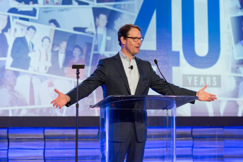 A man speaking at a podium spreads his arms out during a business talk