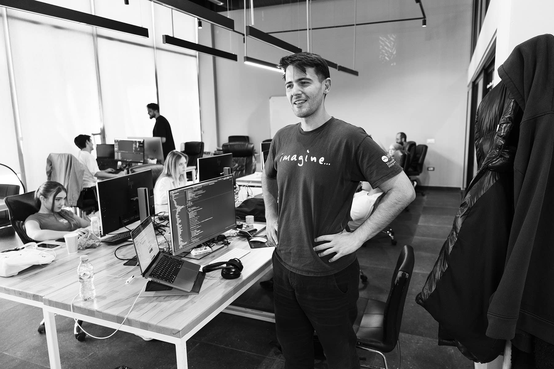 A young man standing next to a computer in an open office space.