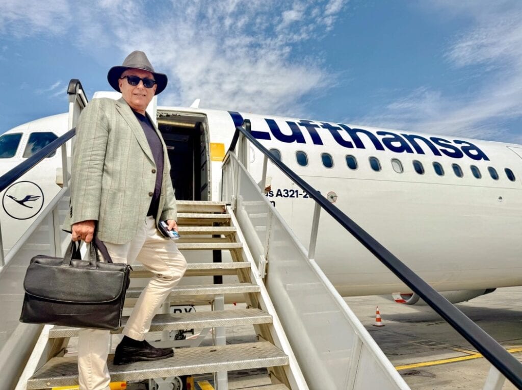 A man wearing a blazer, hat, and carrying a briefcase stands on the steps up to a jet airplane.