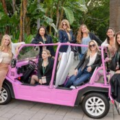 A group of women in a pink golf cart in front of palm trees and other greenery.