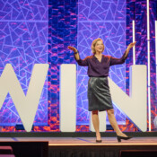 A woman presenting on stage during a business conference, with the word "Win" in the background