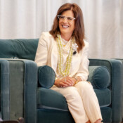 A sharply dressed woman with glasses smiles from an overstuffed chair on stage during a business seminar.