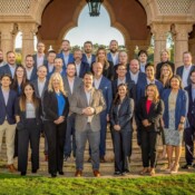 A group of corporate employees in business attire pose outdoors for a group shot in front of architectural pillars.