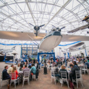 People seated a round tables in a large atrium with an airplane hanging from the ceiling