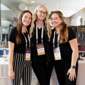 Three women wearing conference badges pose in a hallway.